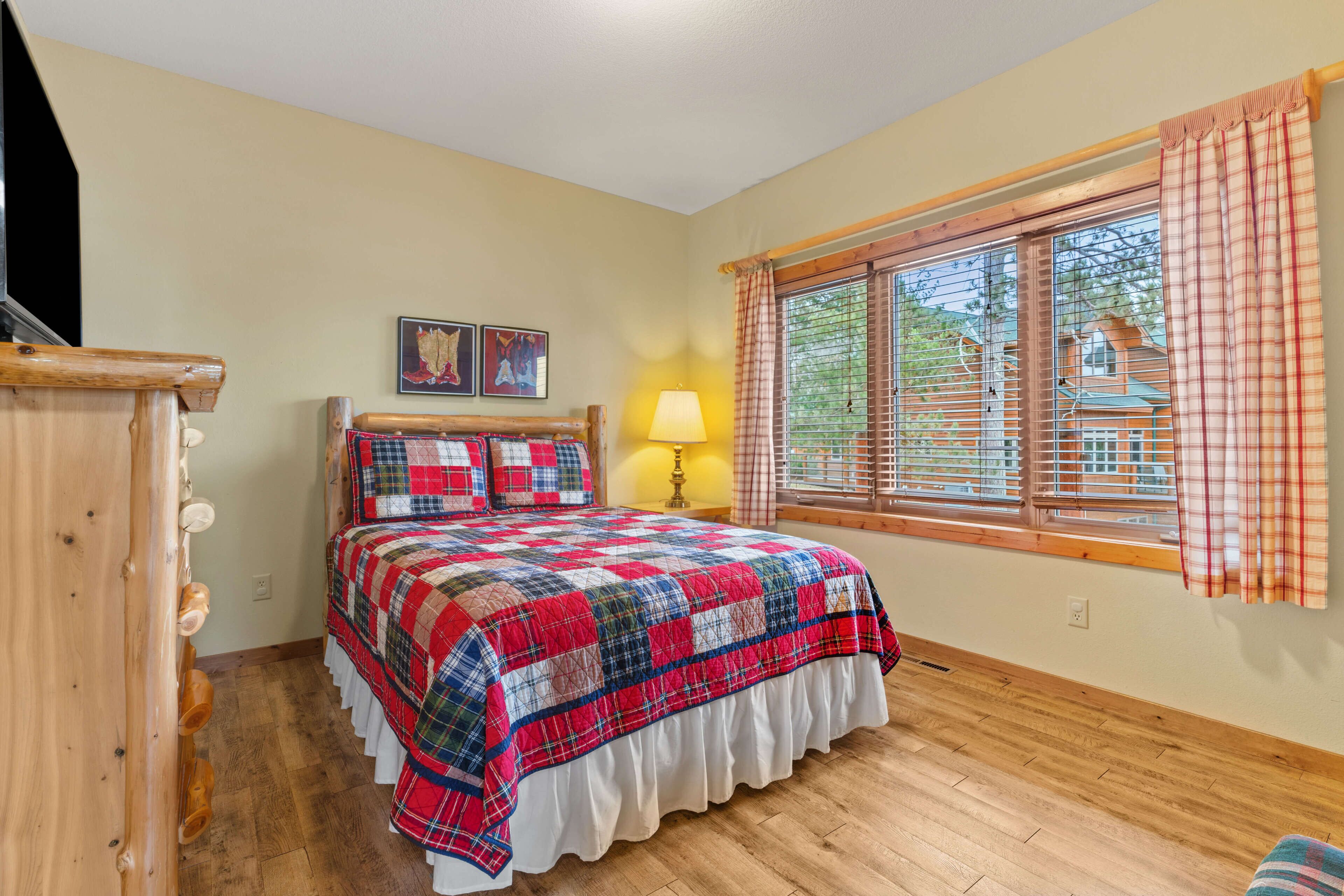 Une chambre avec un plancher en bois, un lit recouvert d’une couverture à carreaux, une table de chevet avec une lampe et une fenêtre avec des stores.