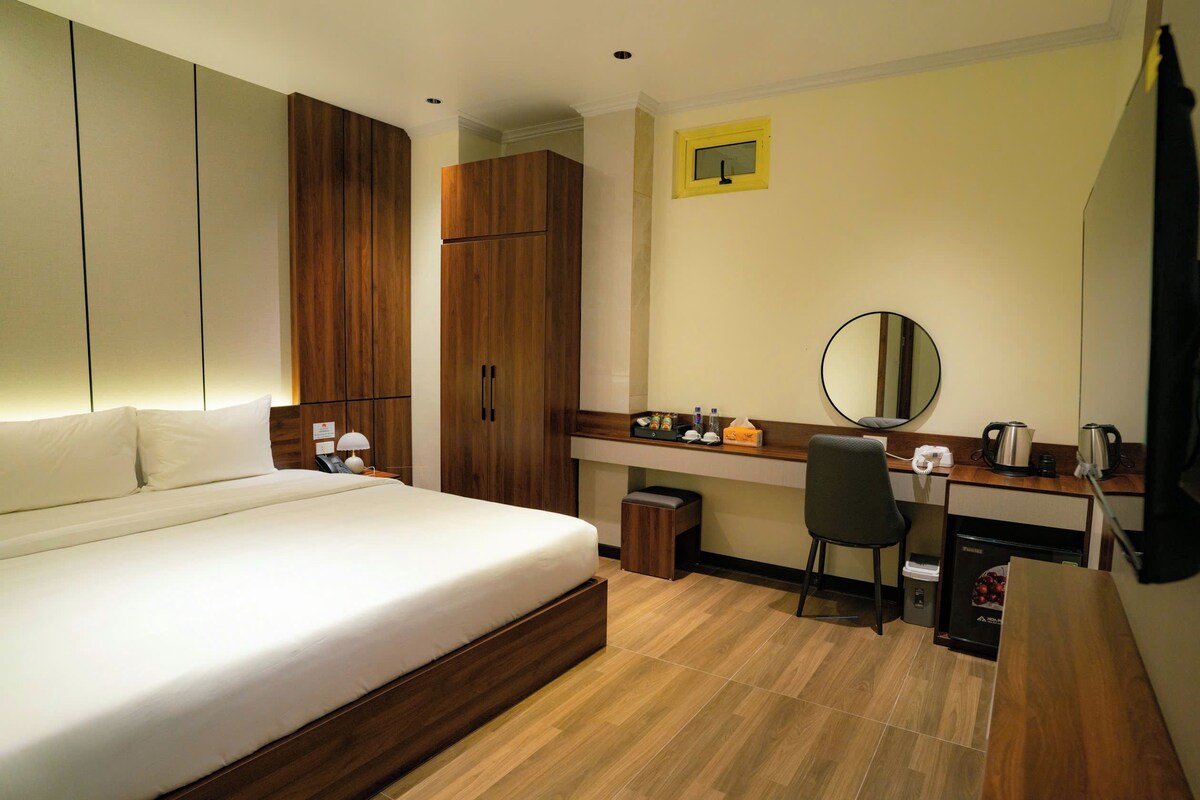 Quarto de hotel com cama, escrivaninha, cadeira, lixeira pequena e espelho.