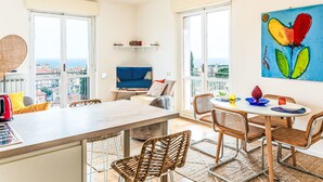 A modern living room with a large window, a dining area, and a view of the city. - Sirio Vista Blu Apartment by Interhome (San Lorenzo al Mare)