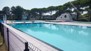 A swimming pool with a diving platform, surrounded by a fence, trees, and a building with a unique architectural design. - Family-friendly villa for eight guests (Bibione)