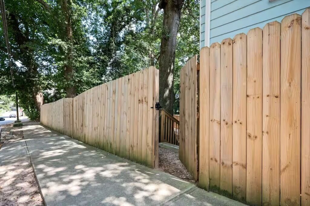 Ein Holzzaun mit einem Tor, ein Betonweg und eine von Bäumen gesäumte Begrenzung.