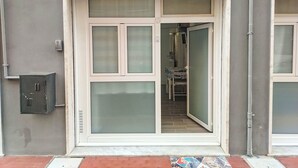 Entrada de edifício com porta de vidro ampla, caixa de utilidades metálica e tapete de entrada ladrilhado.