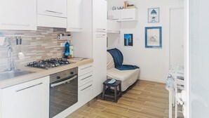 Cozinha compacta com armários brancos, fogão a gás e forno pequeno. Há uma cadeira com almofada azul e um banquinho.