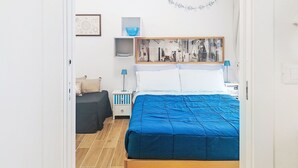 Quarto com cama, mesa de cabeceira, mesinha e abajur azul, além de uma obra de arte emoldurada na parede.