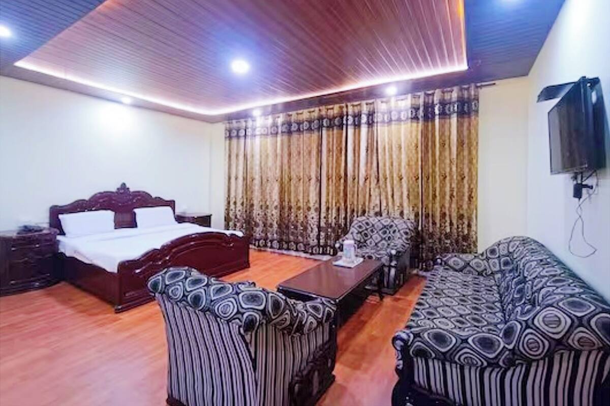 A room with a bed, a sofa, a patterned armchair, a wooden coffee table, and a television on the wall.
