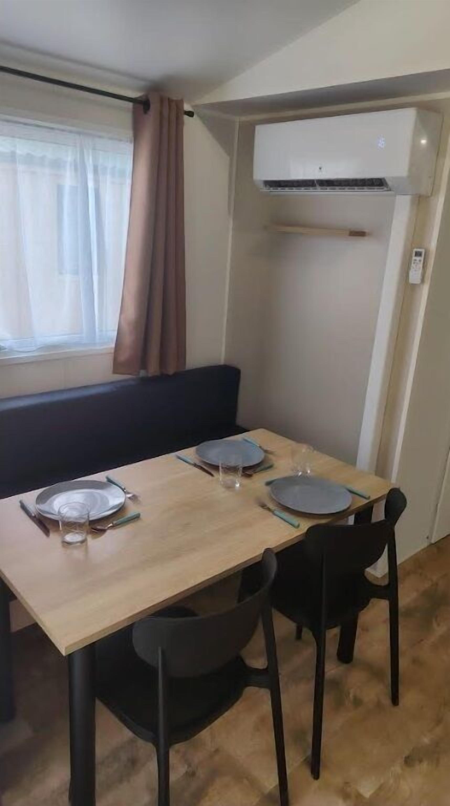 Un área de comedor con una mesa dispuesta para dos personas, una ventana con cortinas y un equipo de aire acondicionado arriba.