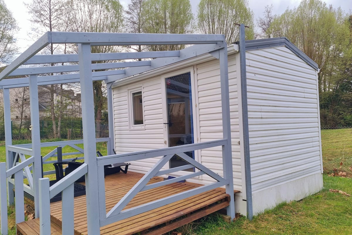 Una cabaña pequeña de un piso con una terraza de madera y un porche con estructura de metal.