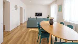 Een moderne woonkamer met een bank, een eettafel met stoelen, een aan de muur bevestigde televisie en een raam met gordijnen.