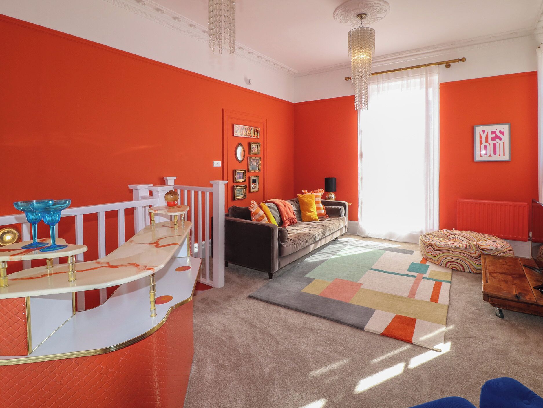 A living room with a red accent wall, a sofa with colorful cushions, a patterned rug, and a white bar area with blue glassware.