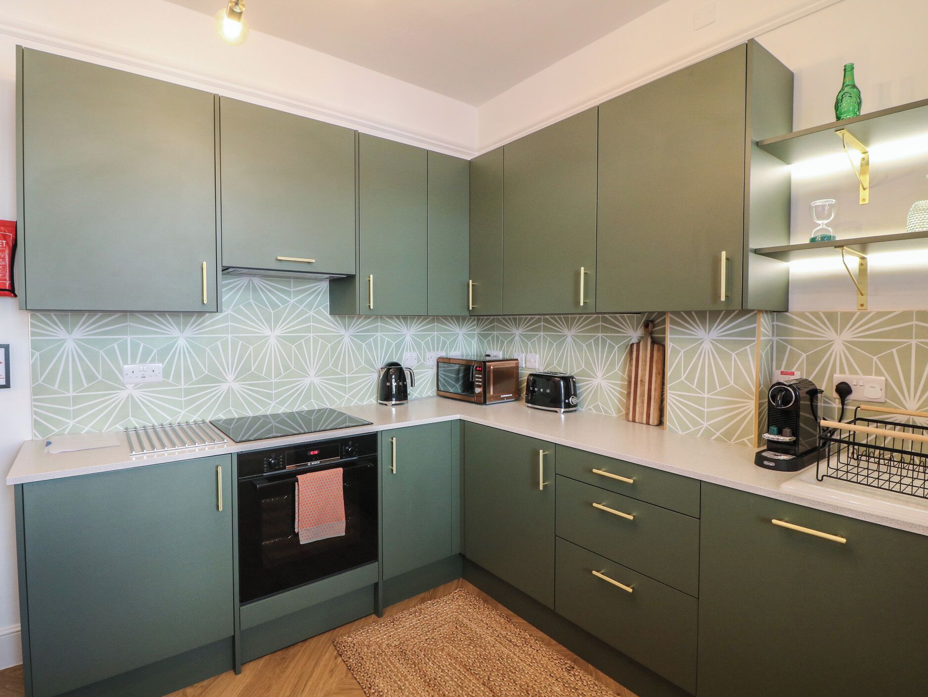 A compact kitchen with a microwave, toaster, and kettle, green cabinets, and a patterned backsplash.