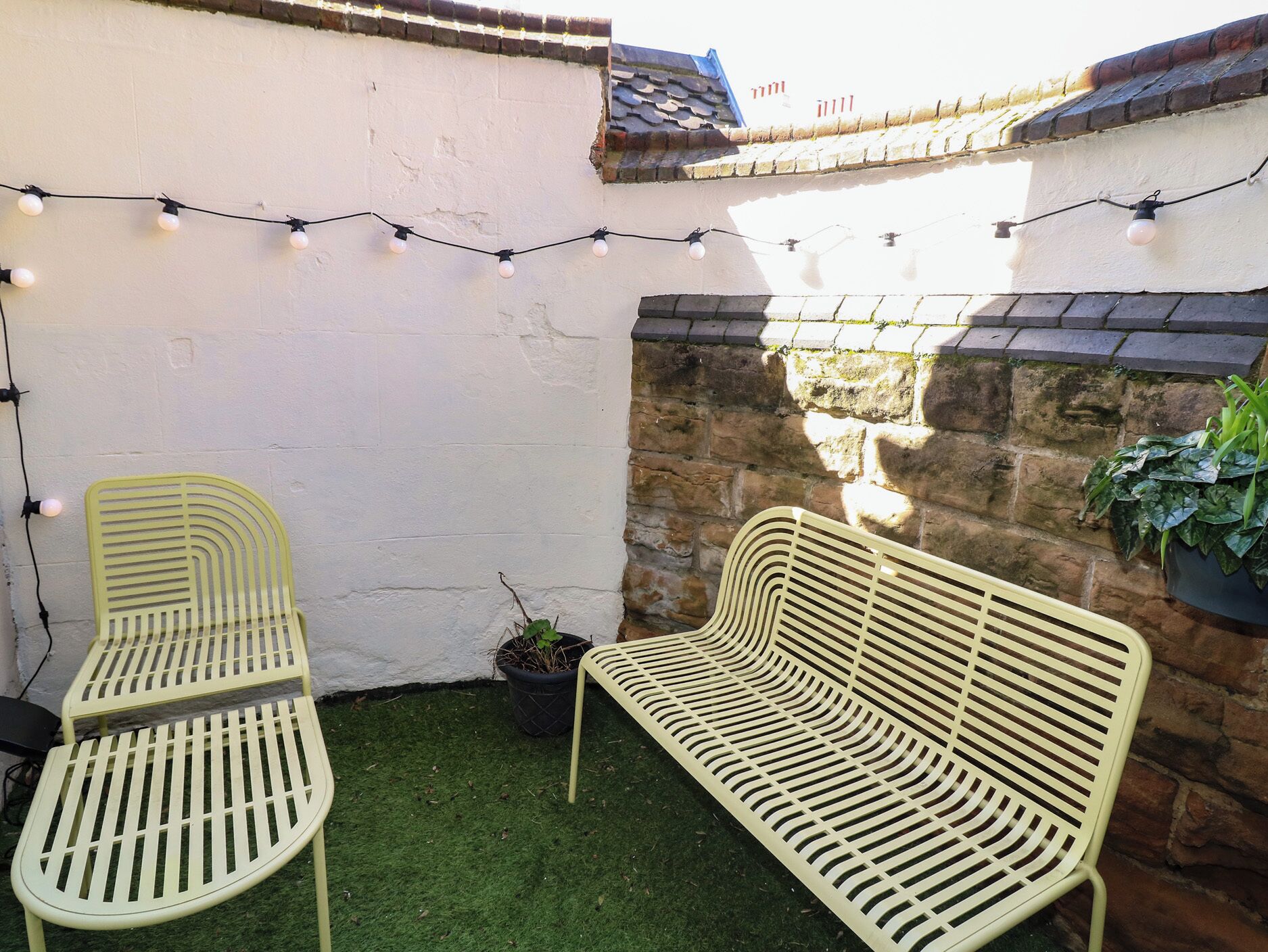 A patio with two metal chairs and a bench, string lights, a potted plant, and a stone wall.