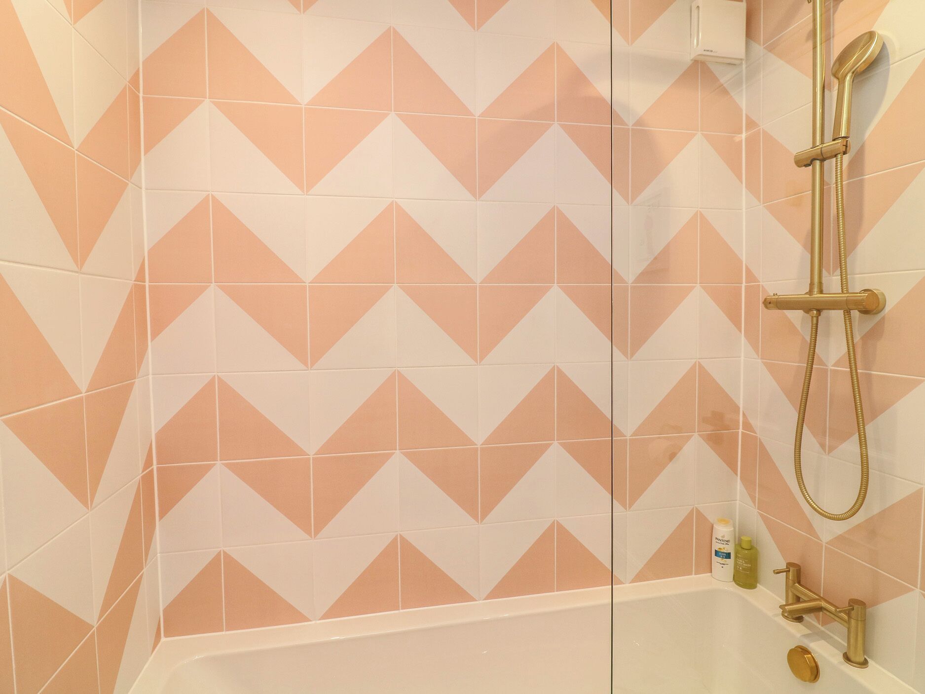 Suihkukaappi, jossa on geometrisesti kuvioituja laattoja, käsisuihkupää ja pullo shampoota.