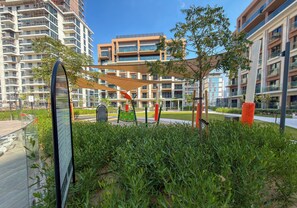 Um parque urbano moderno com uma escultura de vidro, área de recreação infantil e edifícios altos ao fundo.