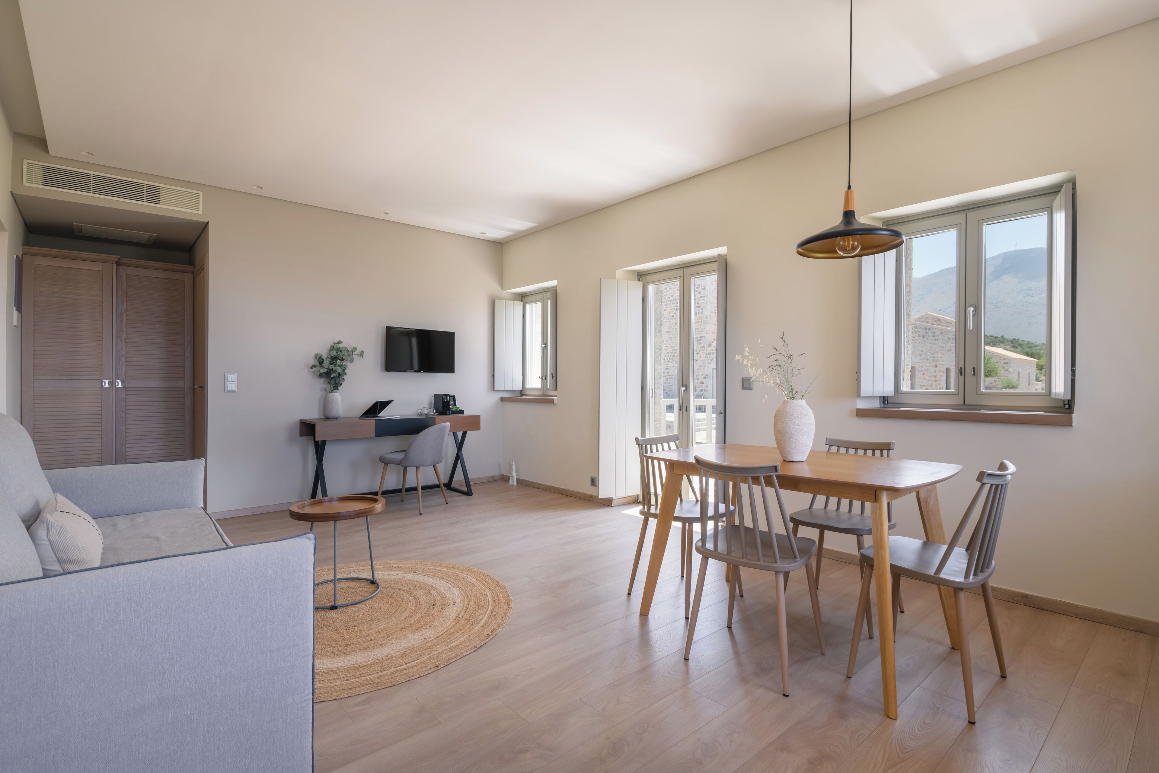 A spacious living area with a dining set, a sofa, a TV mounted on the wall, and large windows offering a view of the hills.