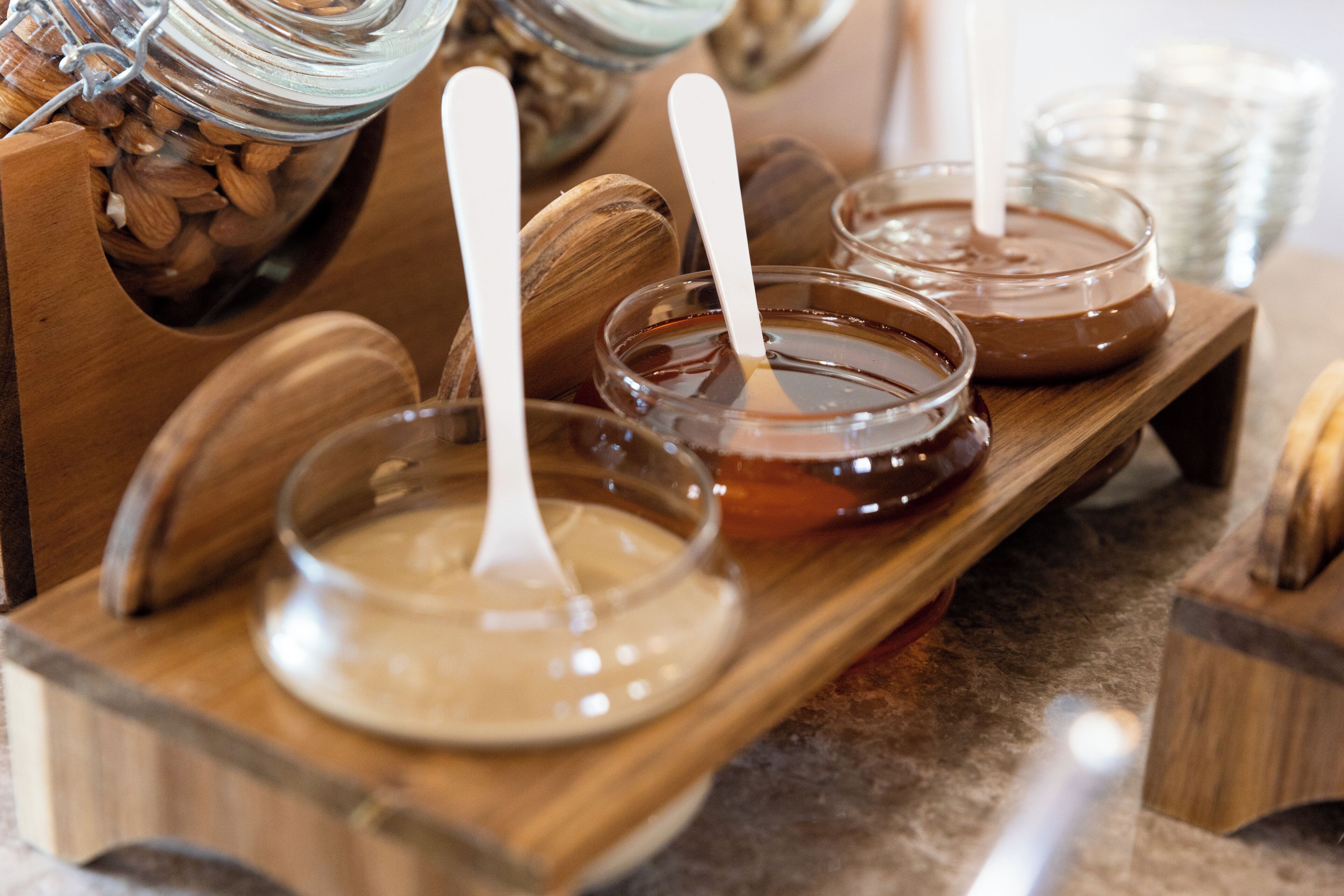 A wooden tray with jars of honey and almonds, and a spoon in each jar.