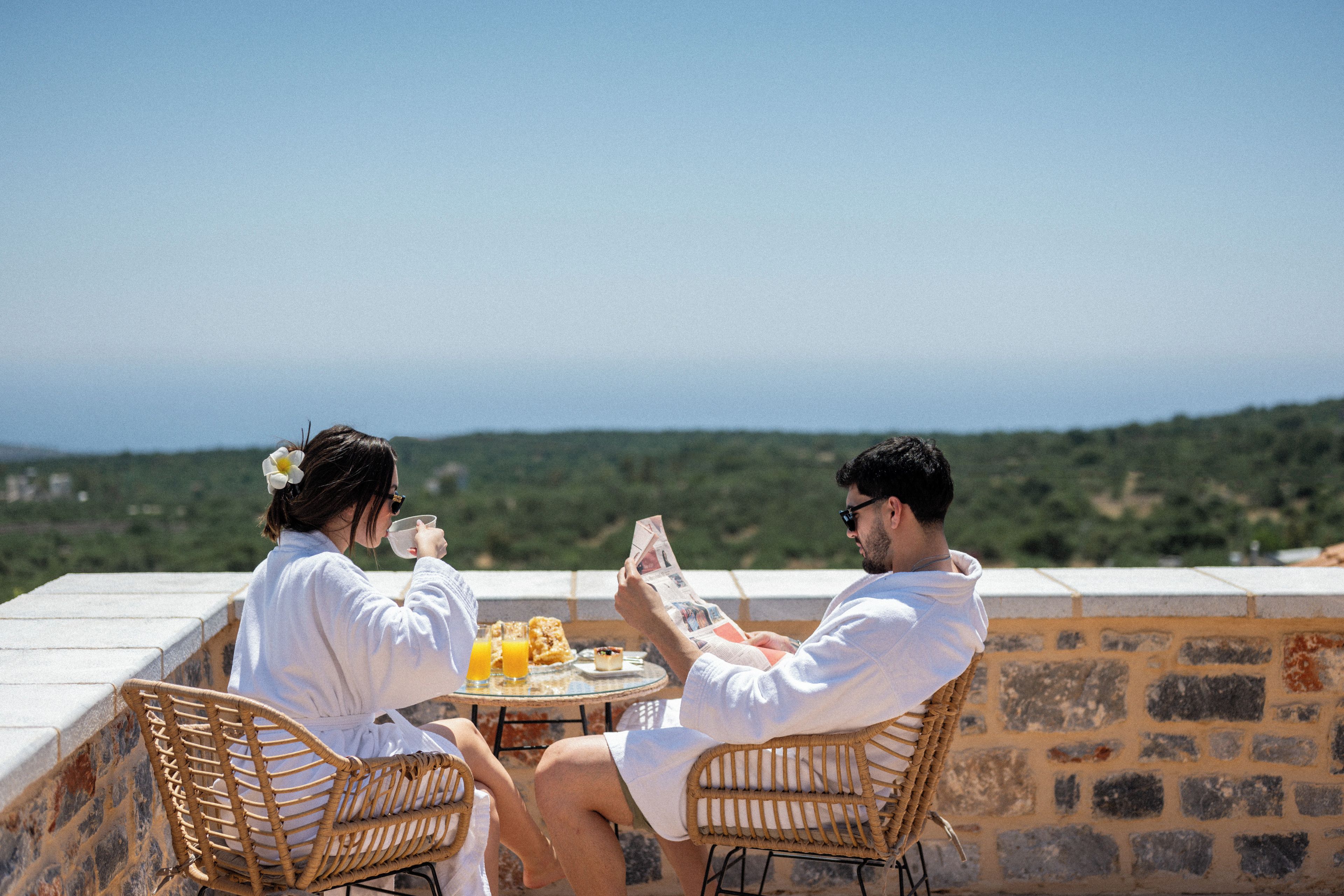 Dos personas sentadas a una mesa con vista al paisaje, un vaso de jugo de naranja y un periódico.