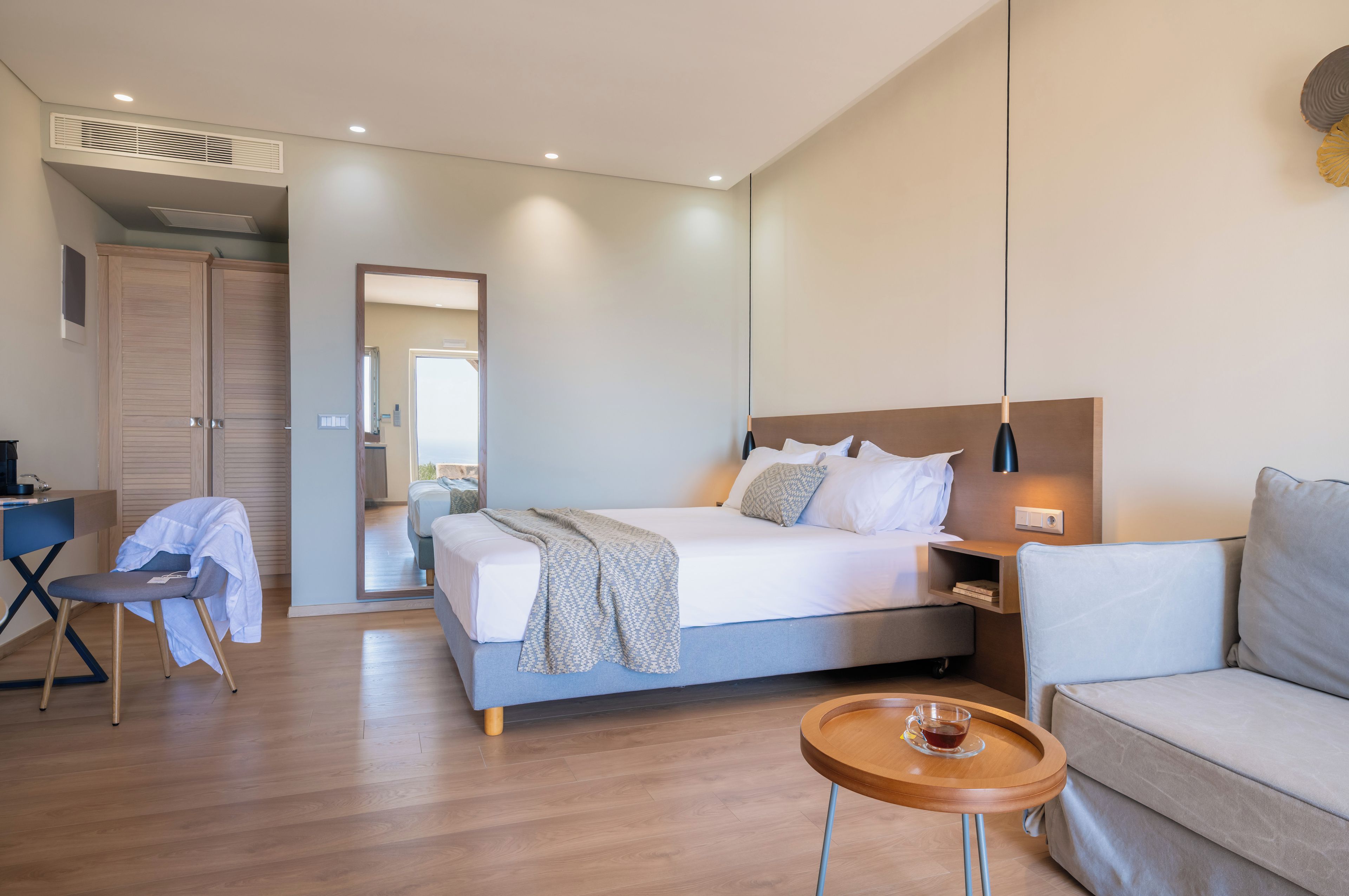 A modern hotel room with a bed, a desk, a chair, a bedside table with a cup, and a sofa.