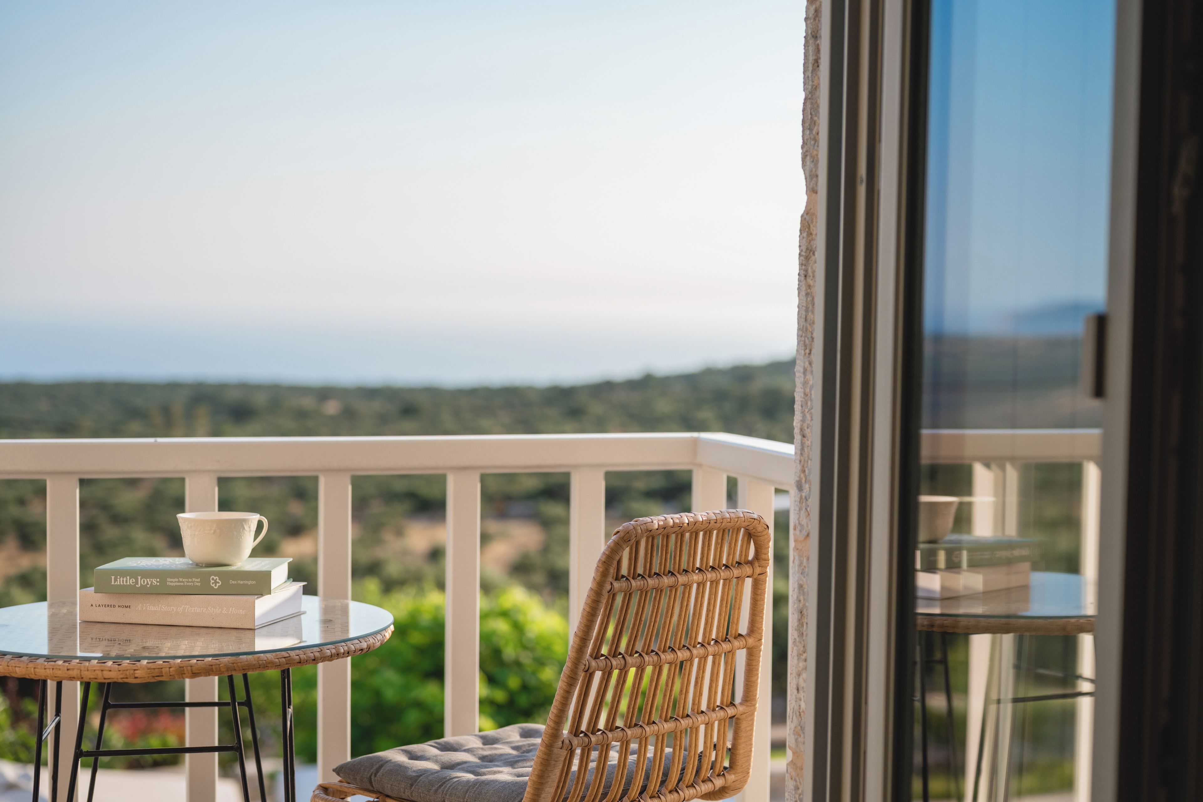 Una silla de mimbre y una mesita redonda con libros y una taza en un balcón con vistas.
