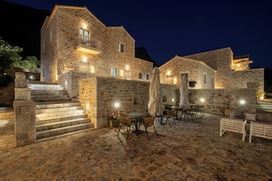 A stone building with a staircase leading to a balcony, outdoor seating area with tables and chairs, and a well-lit entrance.