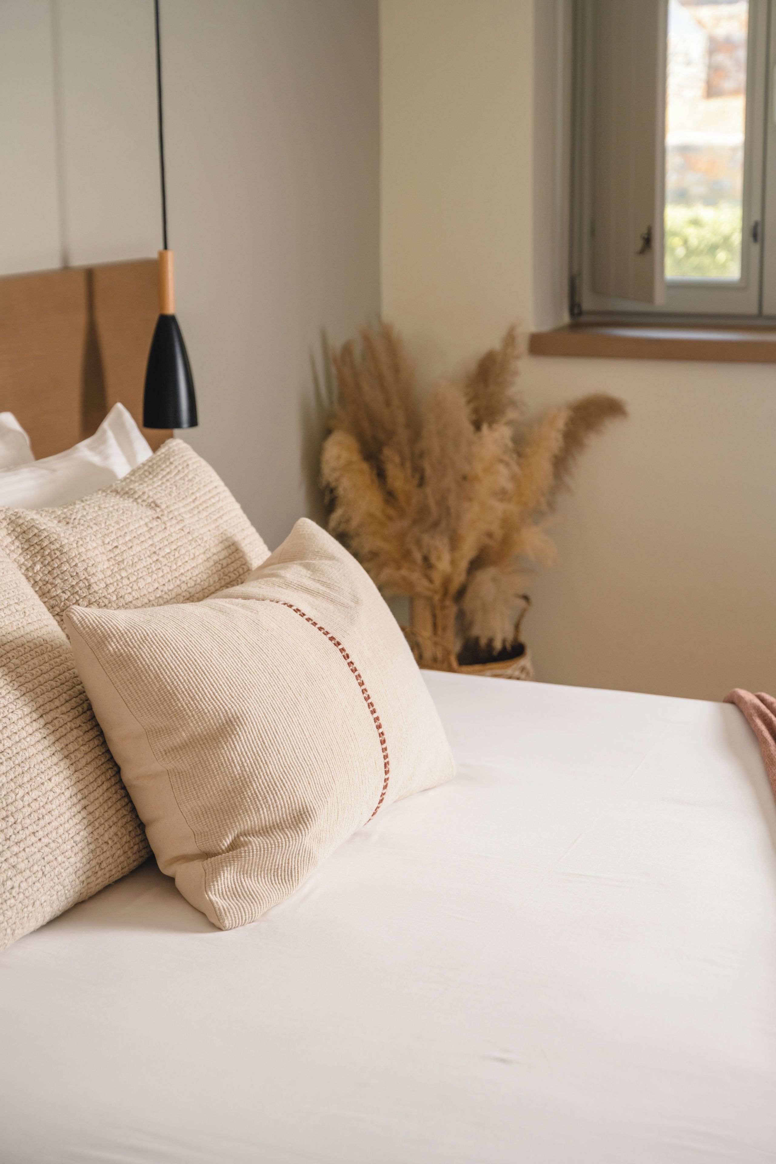 Una cama con ropa de cama blanca y dos almohadas color beige, un cabecero de madera, una lámpara colgante negra y una ventana con vistas a zonas verdes.