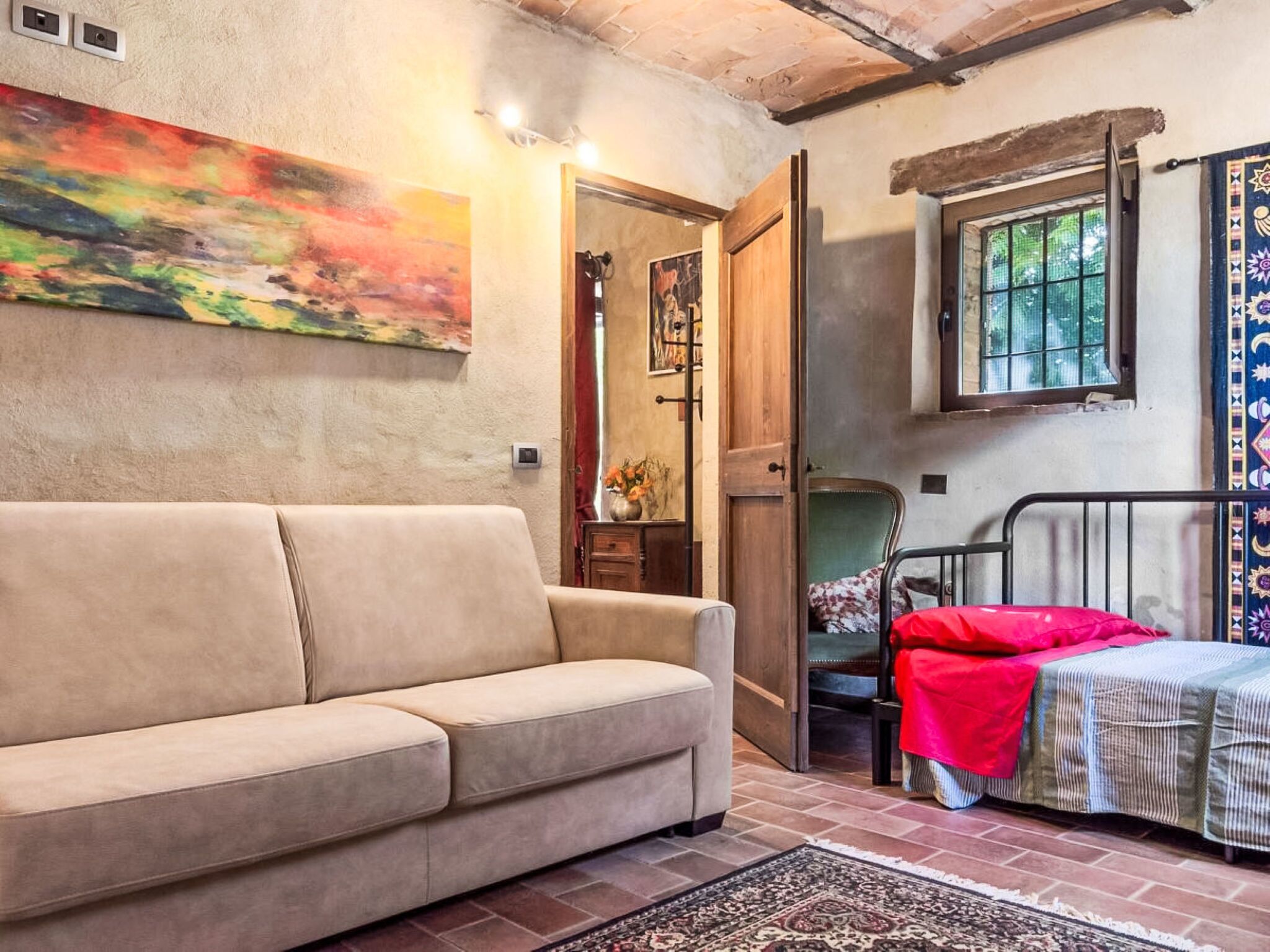 A room with a beige sofa, a bed with a red blanket, a patterned rug, a painting on the wall, and a window with a view.