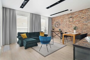 Ein modernes Wohnzimmer mit einem Ecksofa, einem Esstisch mit Stühlen, einem blauen Sessel und einer Backsteinwand.