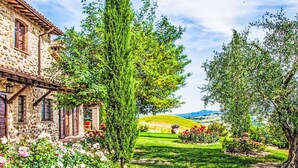 Un bâtiment en pierre avec un jardin, un grand arbre et une vue sur des collines ondoyantes.