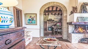 Un intérieur rustique avec une cheminée, un coin repas et une cuisine visible à travers une arche.