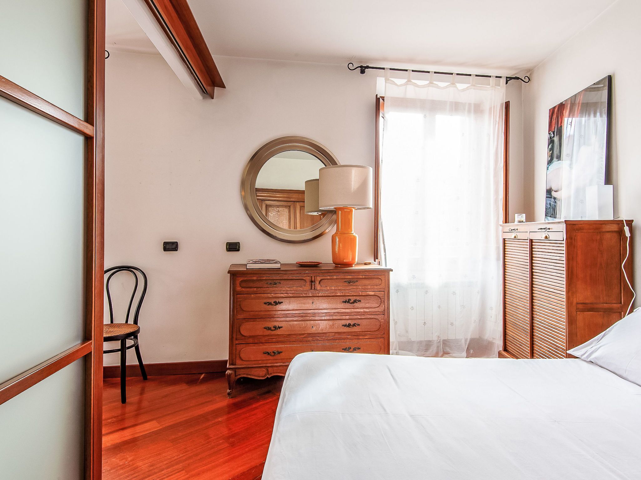 Een slaapkamer met een bed, een dressoir, een stoel, een spiegel en een raam met gordijnen.