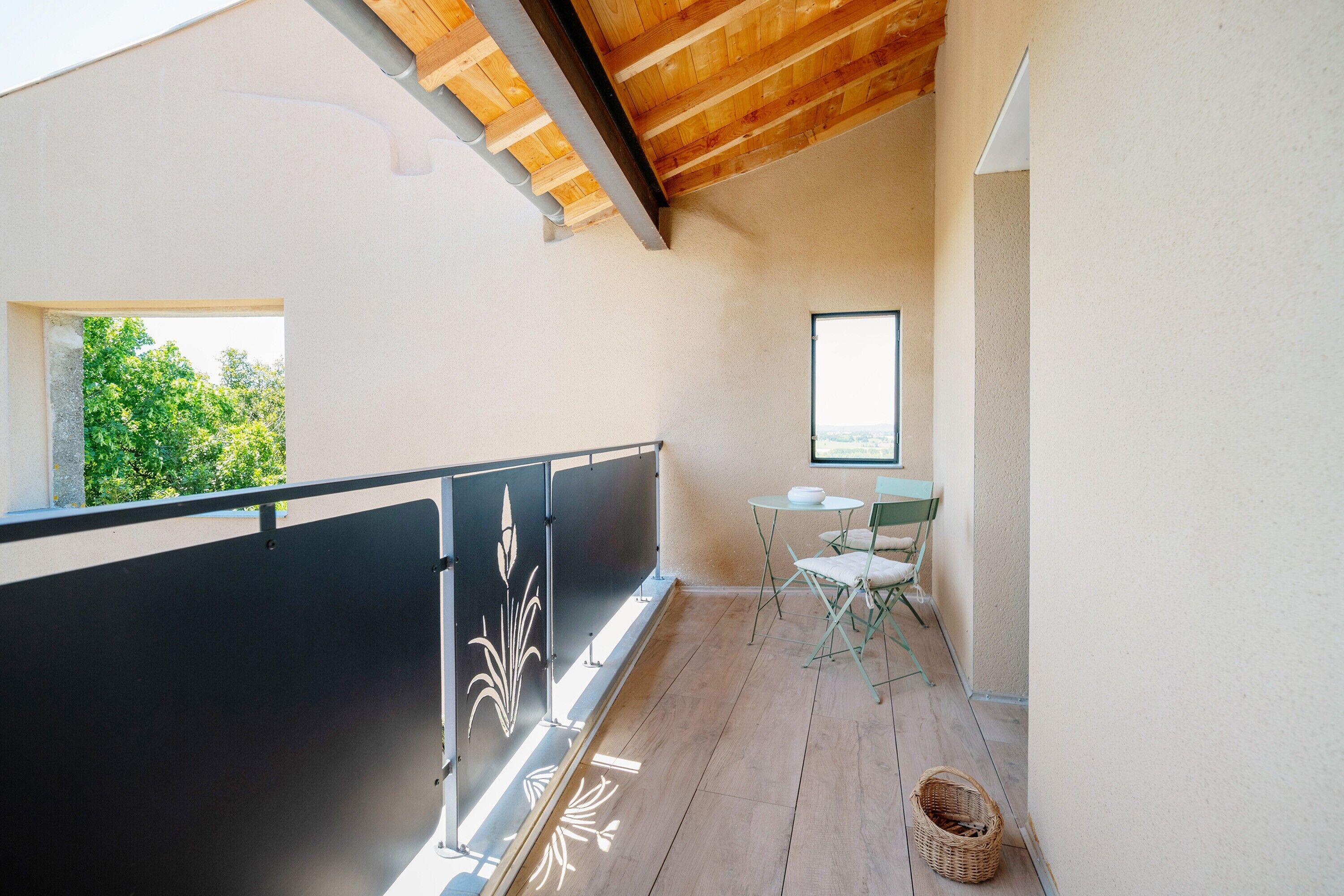 Un balcon doté d’un plafond en bois, d’une rambarde noire agrémentée d’éléments décoratifs, d’une petite table avec des chaises et d’une fenêtre donnant sur un paysage verdoyant.