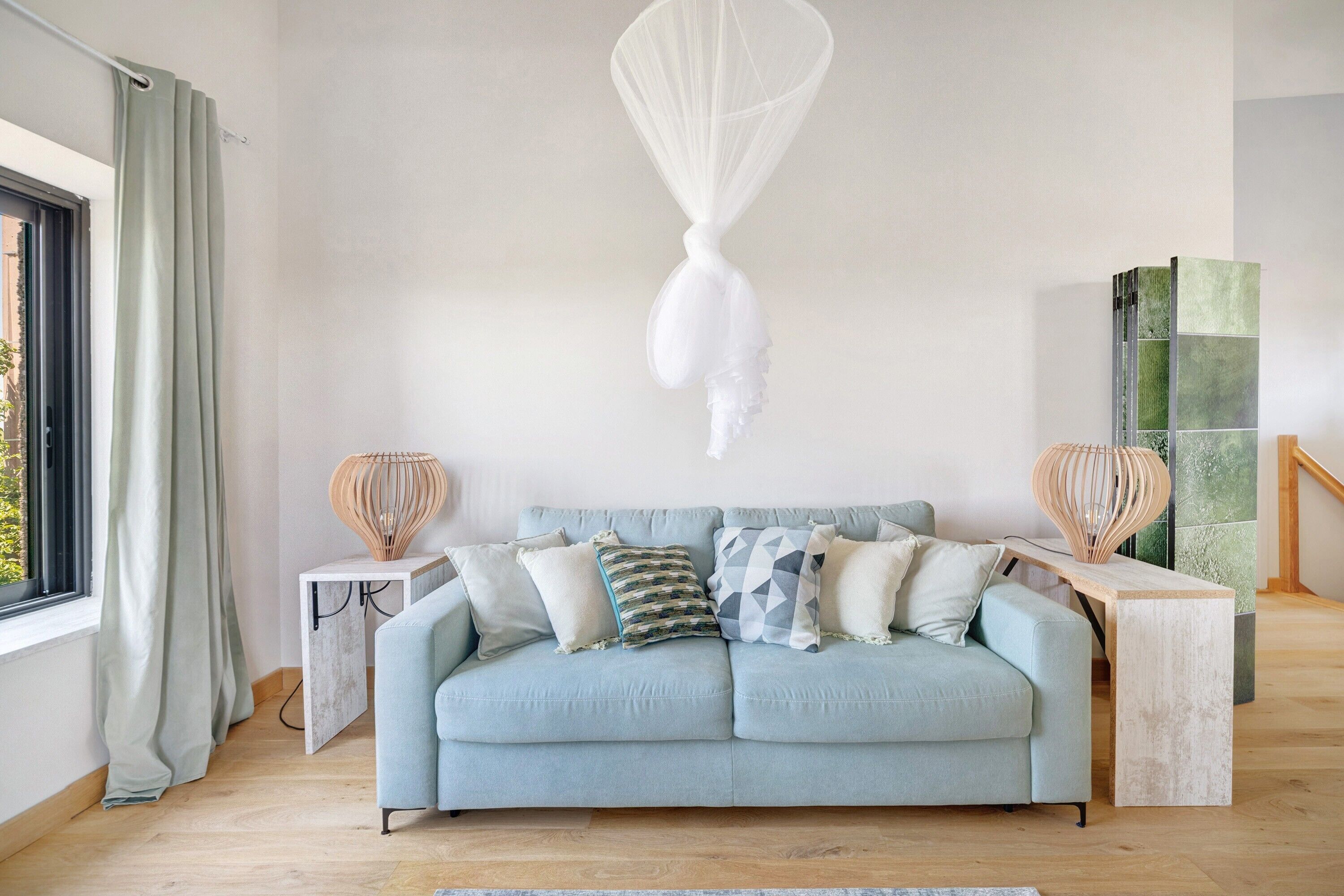 Un sofá azul claro con varios cojines, un objeto colgante blanco y un banco de madera con una lámpara trenzada.