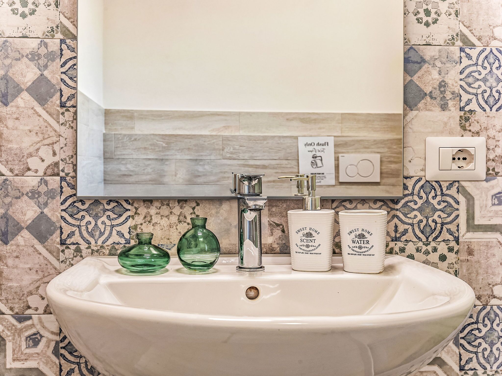 A bathroom with a sink, green glass bottles, and two white containers labeled 'Sweet Home Scent' and 'Sweet Home Water'.