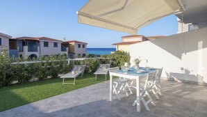 Eine Terrasse mit Tisch und Stühlen, einem weißen Sonnenschirm und Blick auf das Meer.