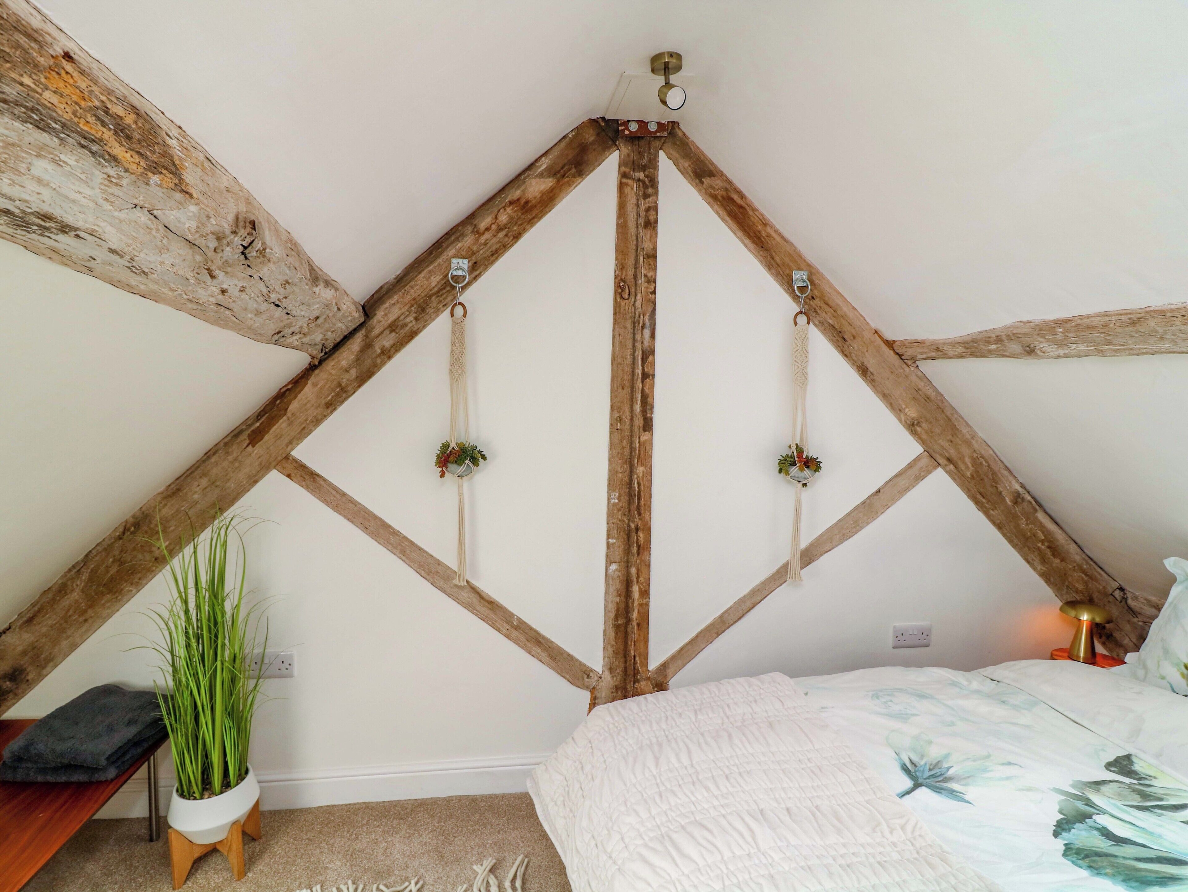 A room with a sloped ceiling, wooden beams, a bed with a floral bedspread, a bedside lamp, a potted plant, and a small wooden stool.