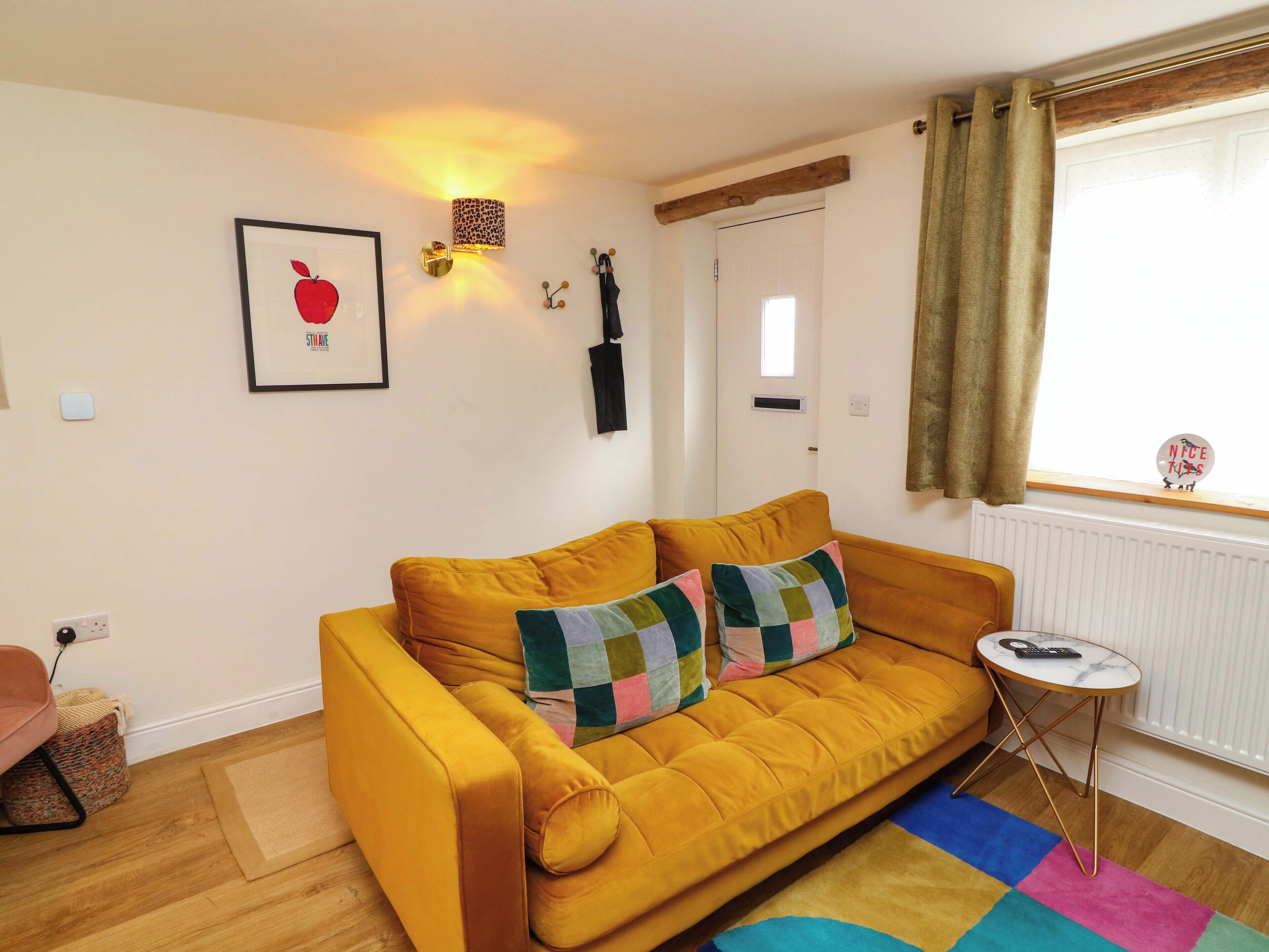 Un sofá amarillo con un cojín de patchwork, una mesita redonda con un control remoto, una obra de arte con forma de manzana en un marco, una lámpara de pared y una ventana con cortinas.