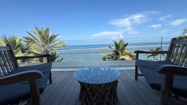 Un terrazzo con un tavolo in vetro e due sedie di legno, con vista su una spiaggia con palme e l'oceano.