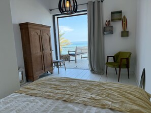 Une chambre à coucher avec un lit, une armoire en bois, une chaise verte, une petite table et une vue sur l’océan par une grande fenêtre.