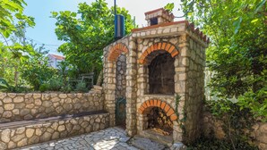 Cocina exterior de piedra con horno de ladrillo, rodeada de follaje y un muro de piedra.
