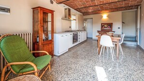 Une cuisine avec des appareils électroménagers blancs, un coin repas avec une table et des chaises, un fauteuil en osier et un radiateur.