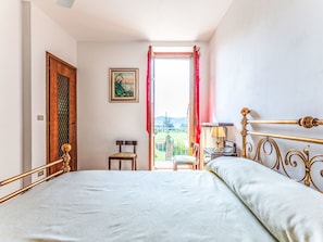 Un dormitorio con cama, una silla, un escritorio, un ventanal con cortinas y un cuadro en la pared.