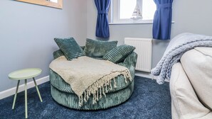 A room with a round green sofa, a small green table, a window with curtains, a radiator, and a bed with a blanket. - Sea Spray (Bideford)