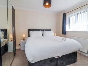 A neatly made bed in a hotel room with a headboard, bedside tables with lamps, a mirror, and a window with curtains.