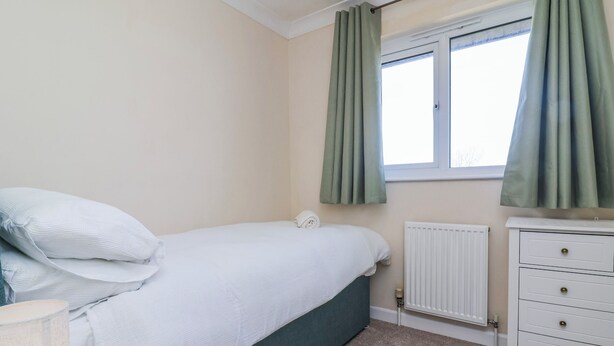 A bedroom with a bed, a dresser, a window with curtains, and a radiator. - Sea Spray (Bideford)
