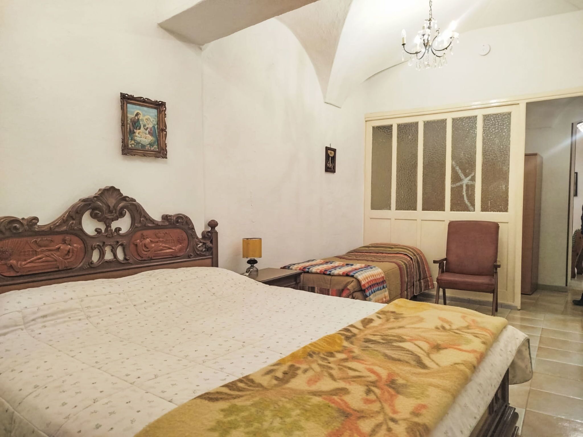 Une chambre à coucher avec un grand lit, un plus petit lit, une chaise, une lampe et une fenêtre avec du verre dépoli.