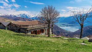 Ein Steinhaus mit geneigtem Dach, einer Holzterrasse und Blick auf einen See und Berge.