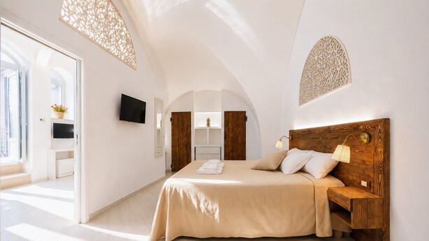 A bedroom with a large bed, wooden headboard, a TV on the wall, and a view of the outside. - Casa Marosa – In the Heart of the Village (Monte Sant'Angelo)