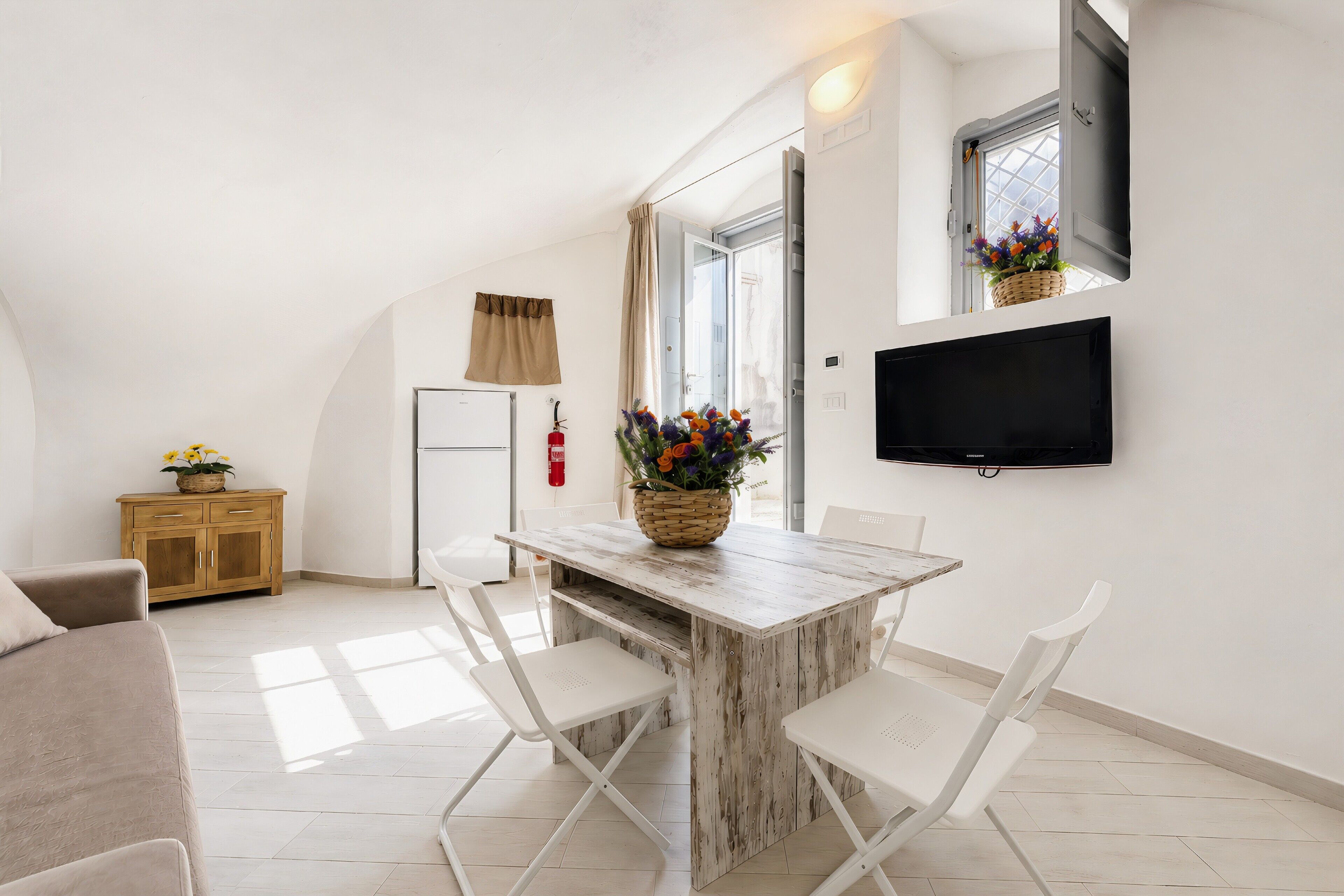 Una cucina moderna con un tavolo di legno e sedie bianche, una televisione appesa al muro e una finestra con un cesto di fiori.