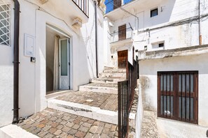 A narrow alleyway with a set of stone steps leading to a door, flanked by white buildings with balconies and a brown gate.