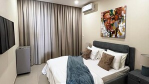 A modern hotel room with a large bed, a flat-screen TV, an air conditioning unit, and a painting on the wall. - CMH SARL (Mbour)