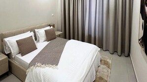 A modern hotel room with a bed, a nightstand, a lamp, a large window with curtains, and an air conditioning unit. - CMH SARL (Mbour)